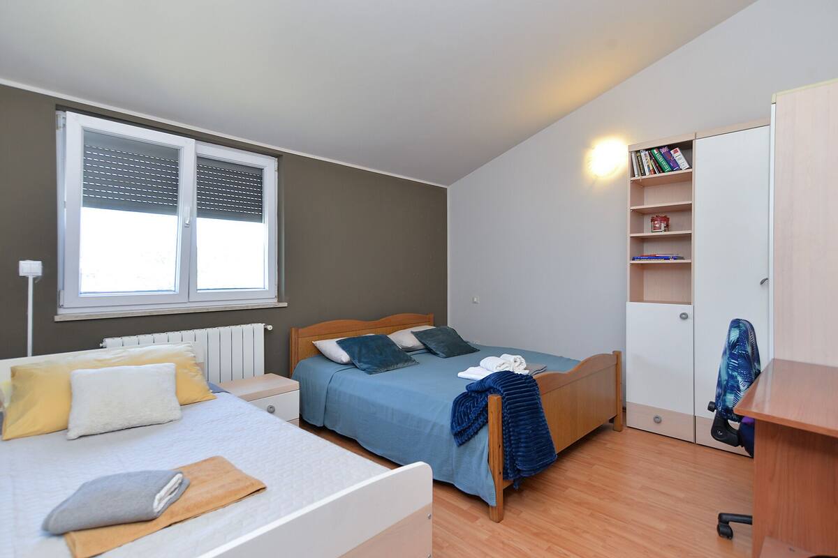Une chambre à coucher comprenant un lit, une table de chevet, un radiateur, une fenêtre avec des stores, une bibliothèque et une armoire.