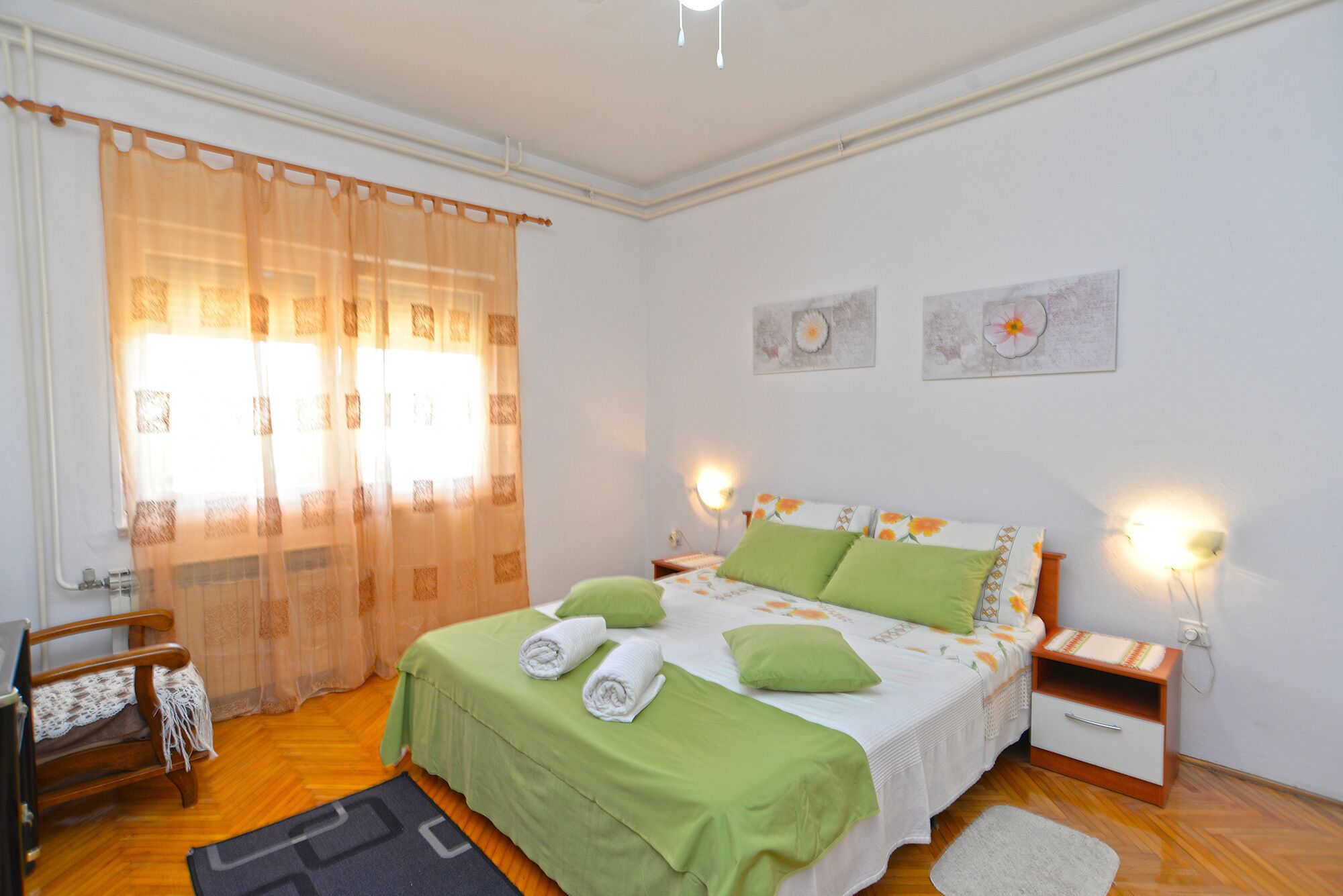 Quarto com cama, almofadas verdes, mesa de cabeceira, cadeira de madeira e janela com cortinas translúcidas.