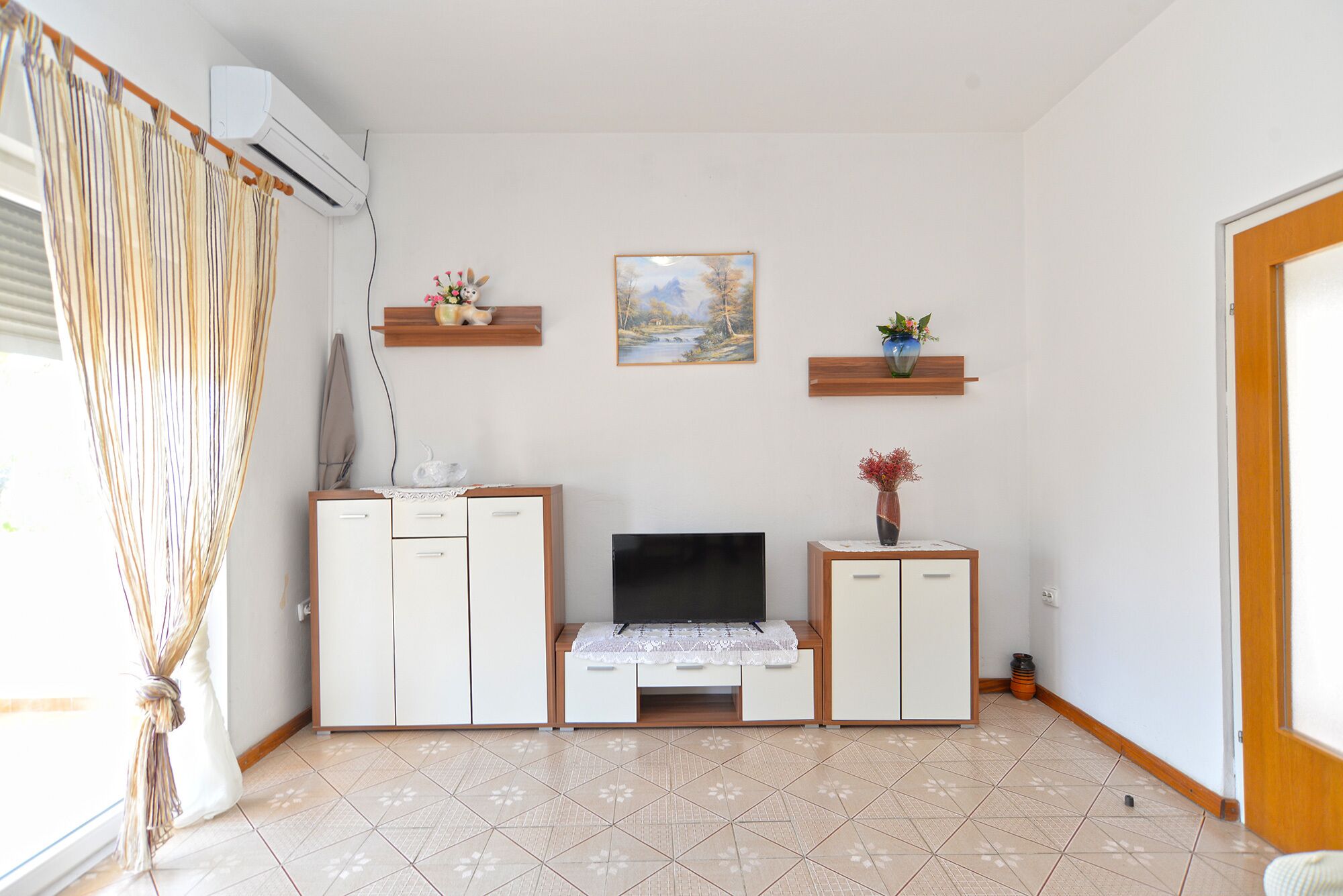 Ein kompakter Wohnbereich mit Fernseher, zwei Schränken, einem kleinen Tisch und einem Fenster mit Vorhängen.
