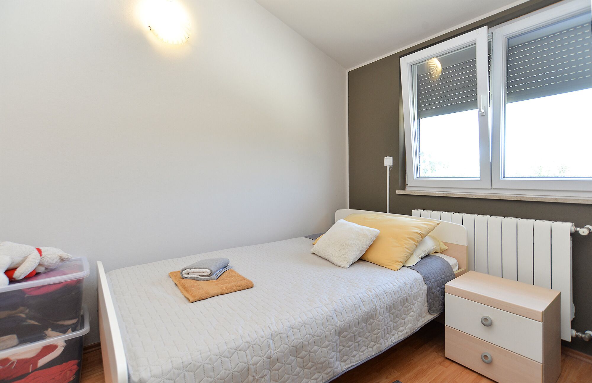 Een kleine, verzorgde slaapkamer met een enkel bed, een nachtkastje, en een raam met jaloezieën.