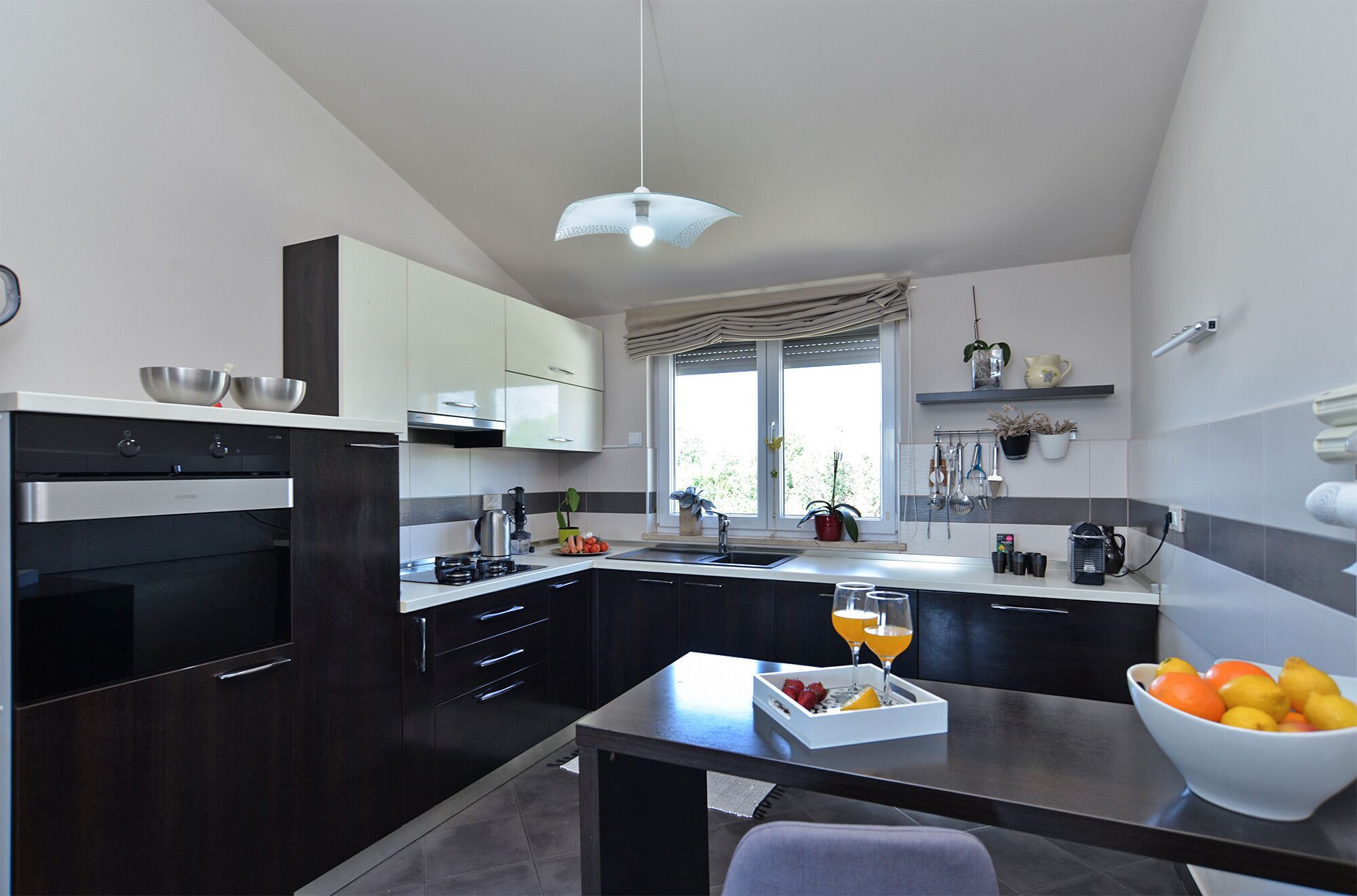 A modern kitchen with dark cabinetry, a built-in oven, a window with a view, and a dining area with a table and chairs.