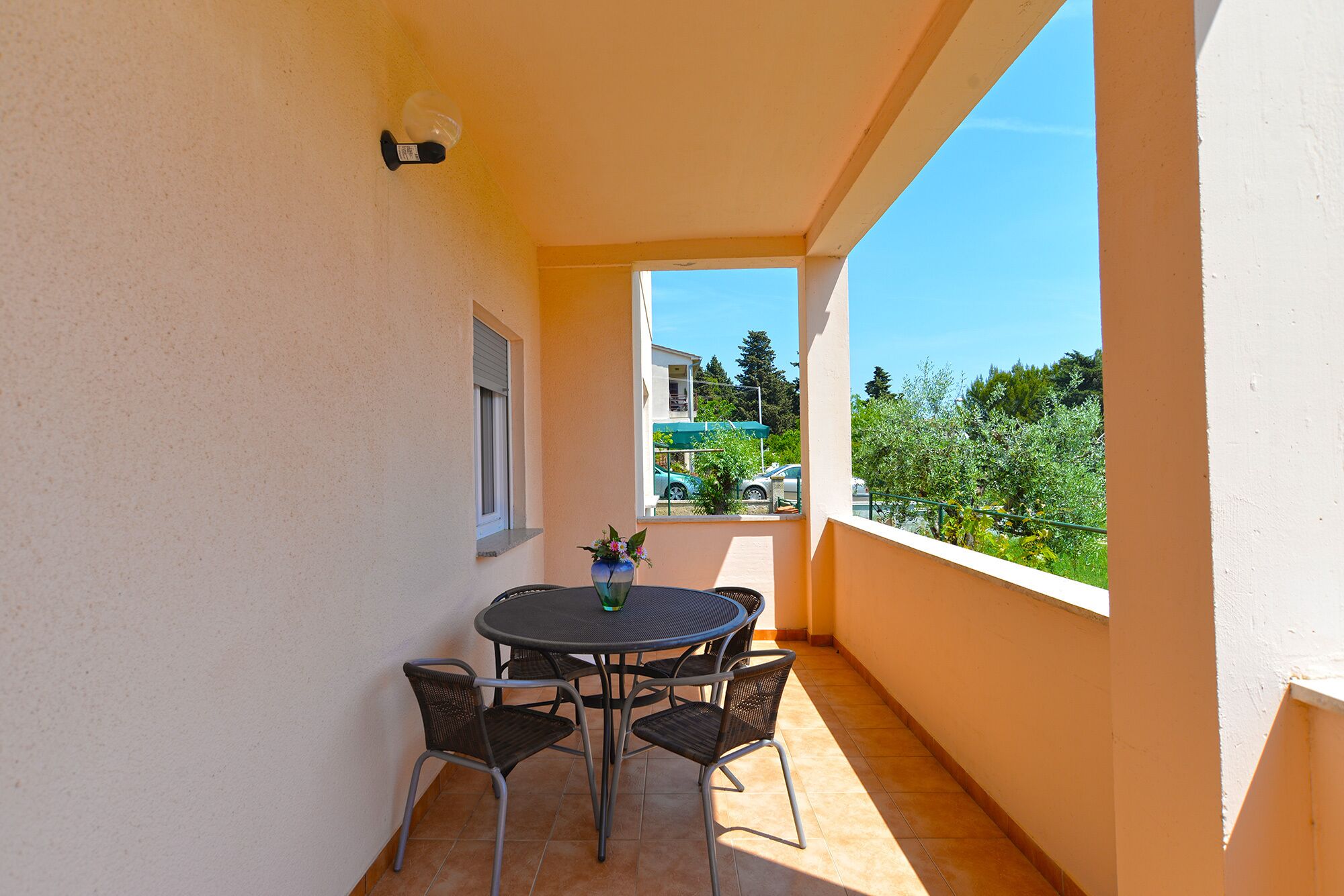 A balcony with a round table and four chairs, a potted plant, and a view of parked cars and greenery.