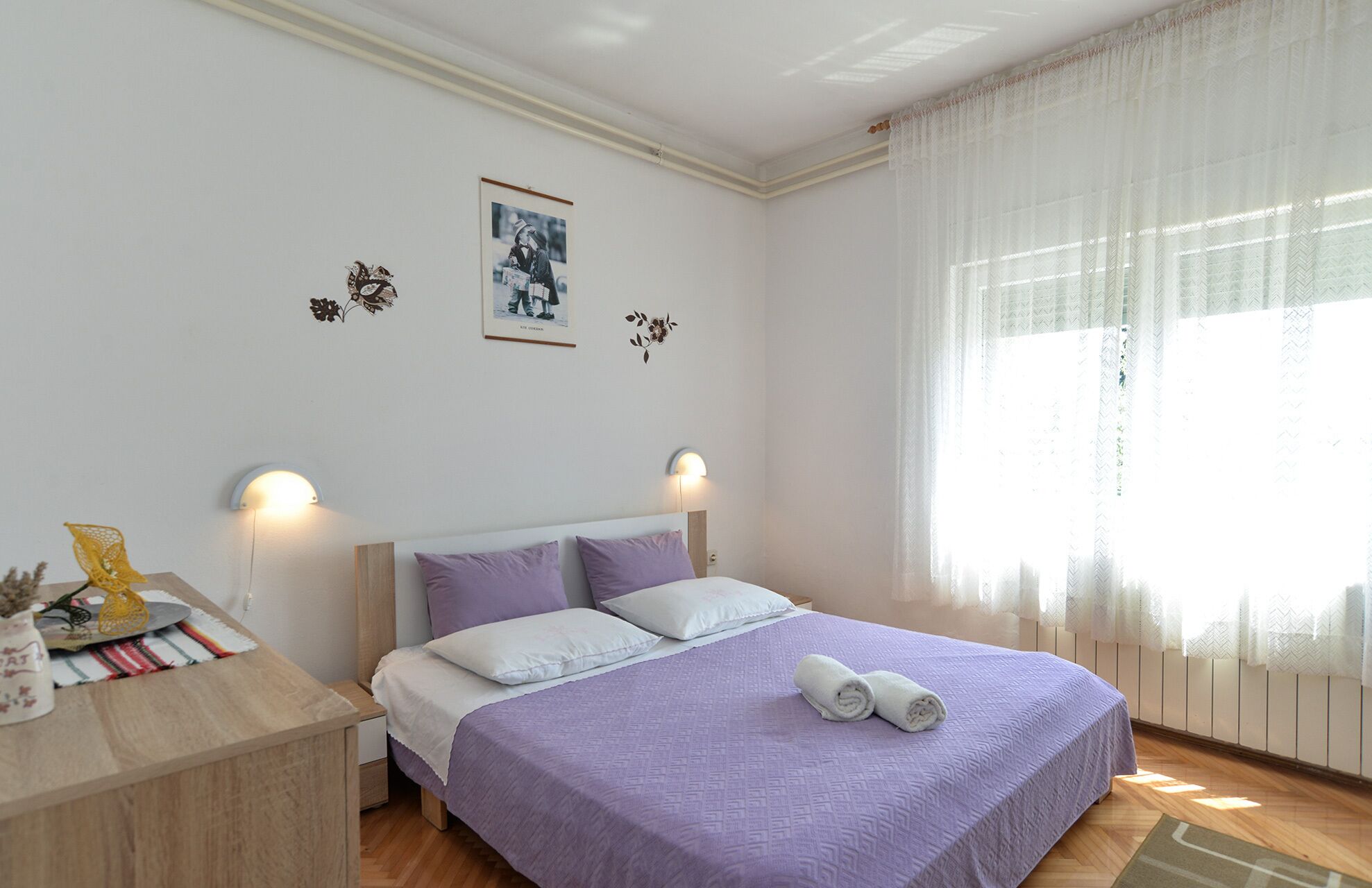 Un dormitorio con una cama, dos lámparas de pared, un cuadro enmarcado y una ventana con cortinas translúcidas.