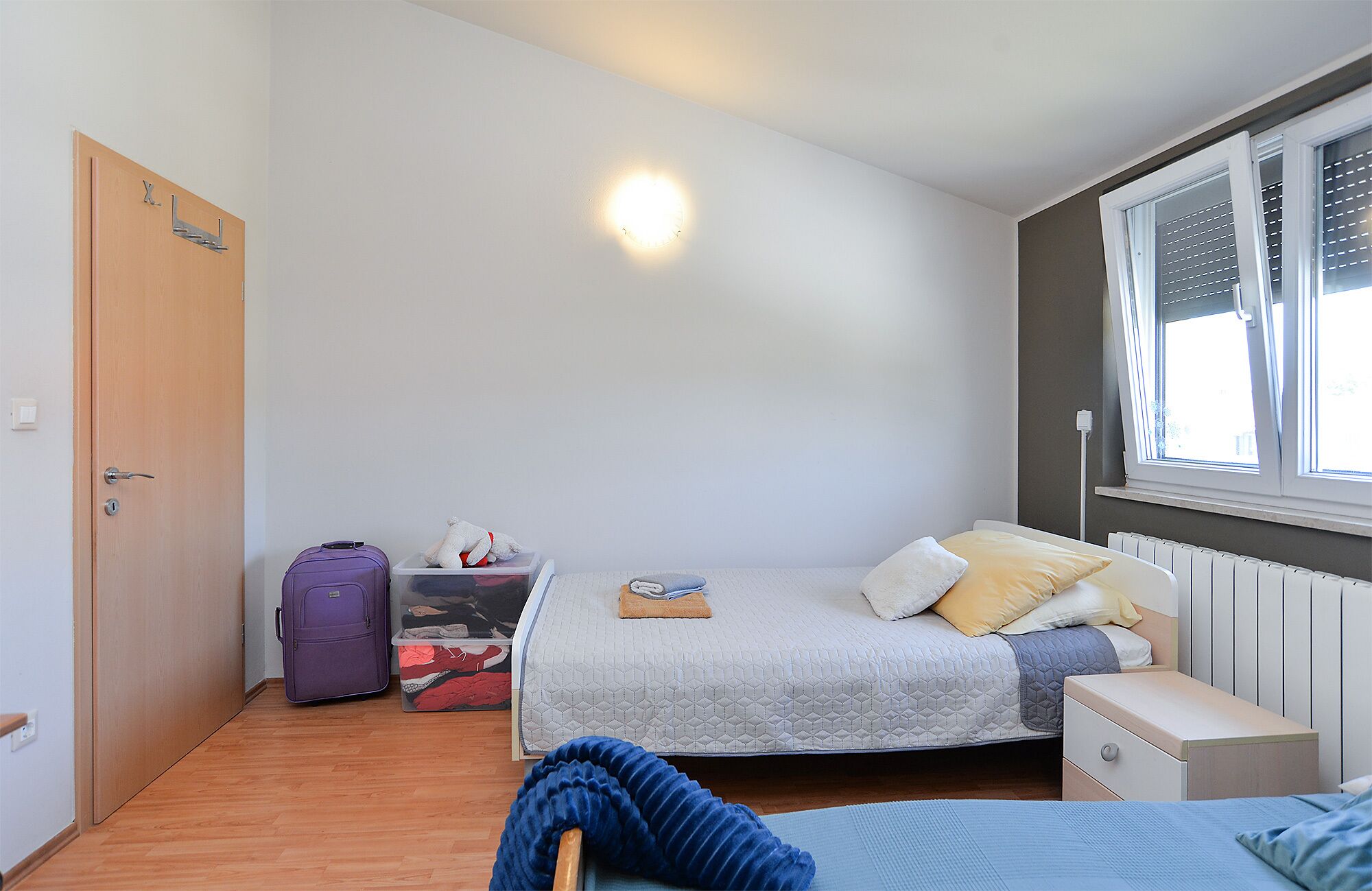 A bedroom with a bed, a nightstand, a window with blinds, a suitcase, and a storage box.