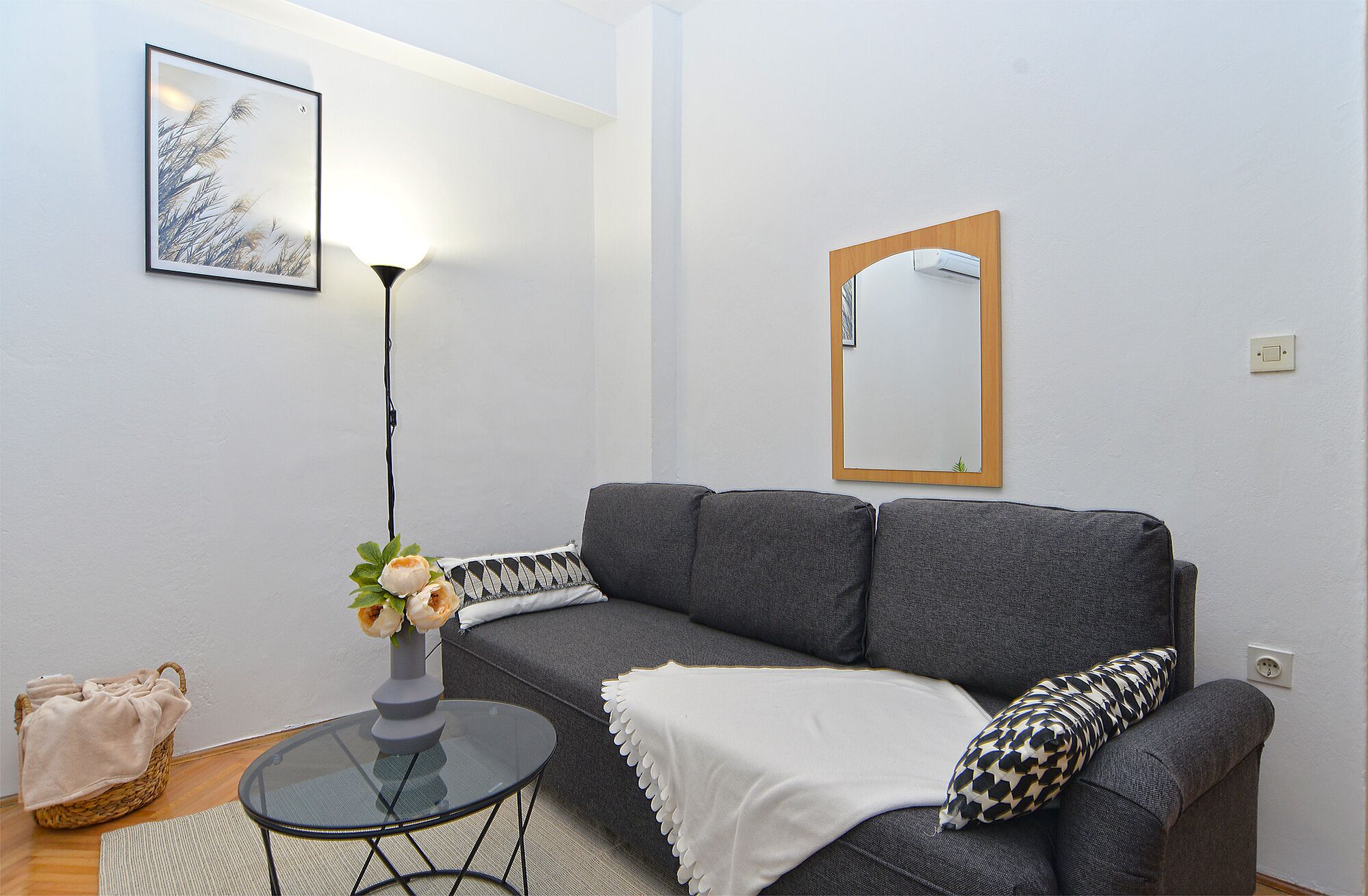 Een woonkamer met een grijze bank, een ronde glazen salontafel, een vloerlamp, een lijst met een afbeelding, een spiegel en een mand met een plaid.