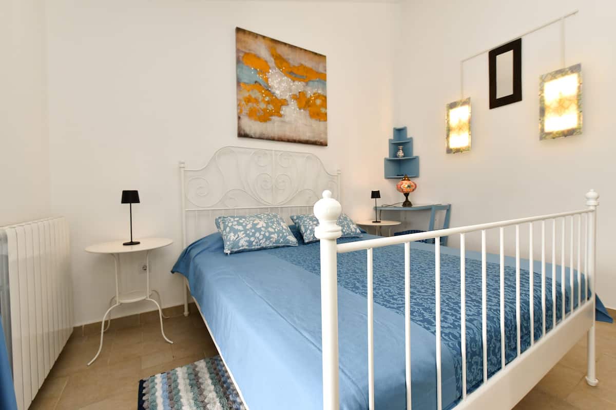 Ein Schlafzimmer mit weißem Metallbettgestell, blauer Bettdecke und passenden Kissen. Ein weißer Heizkörper, ein kleiner runder Tisch mit Lampe und ein Globus auf einem Nachttisch sind ebenfalls vorhanden. Über dem Bett hängt ein Gemälde.