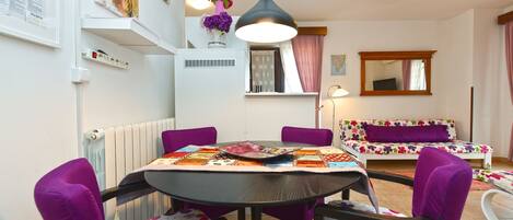 Een eetruimte met een ronde tafel, paarse stoelen en een tafelkleed met bloemen.