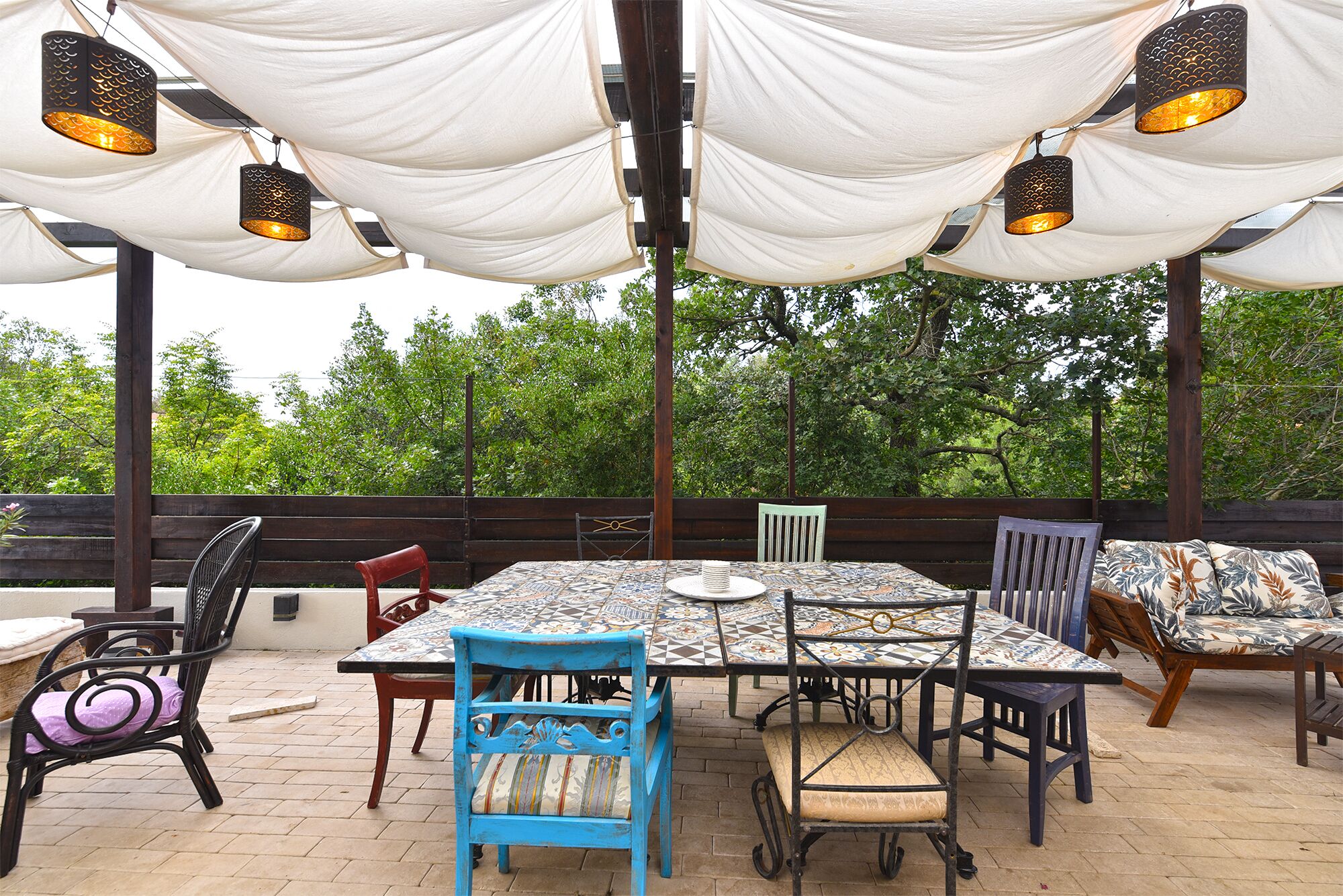 An outdoor patio with a tiled table and chairs, draped white fabric overhead, and a wooden fence in the background.