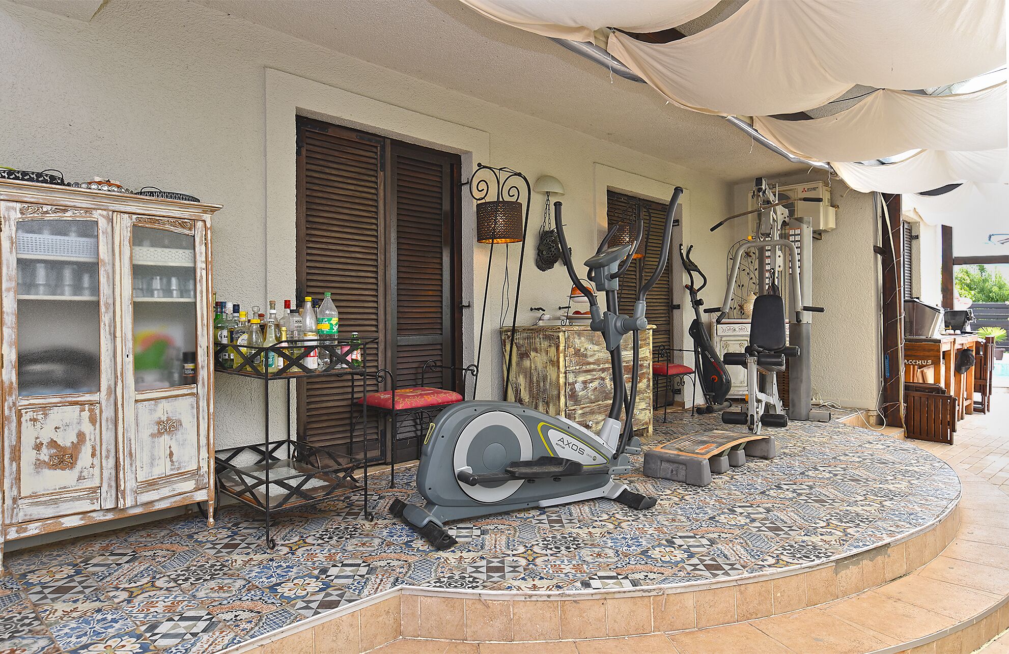 A patio with exercise equipment, a cabinet with bottles, and a tiled floor.