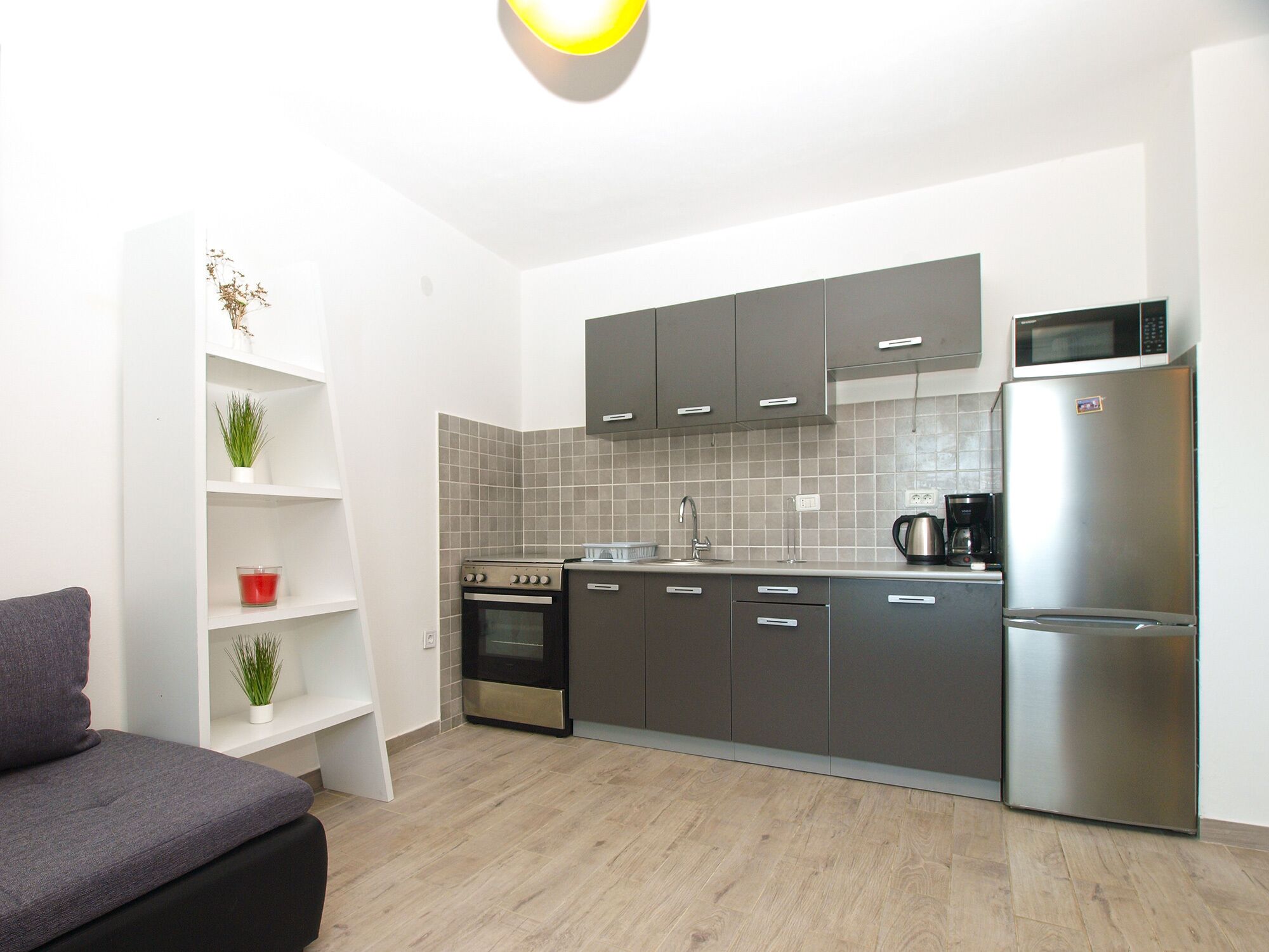 A compact kitchen with grey cabinets, a stainless steel refrigerator, and a microwave. There's a small shelf with plants and a red candle.