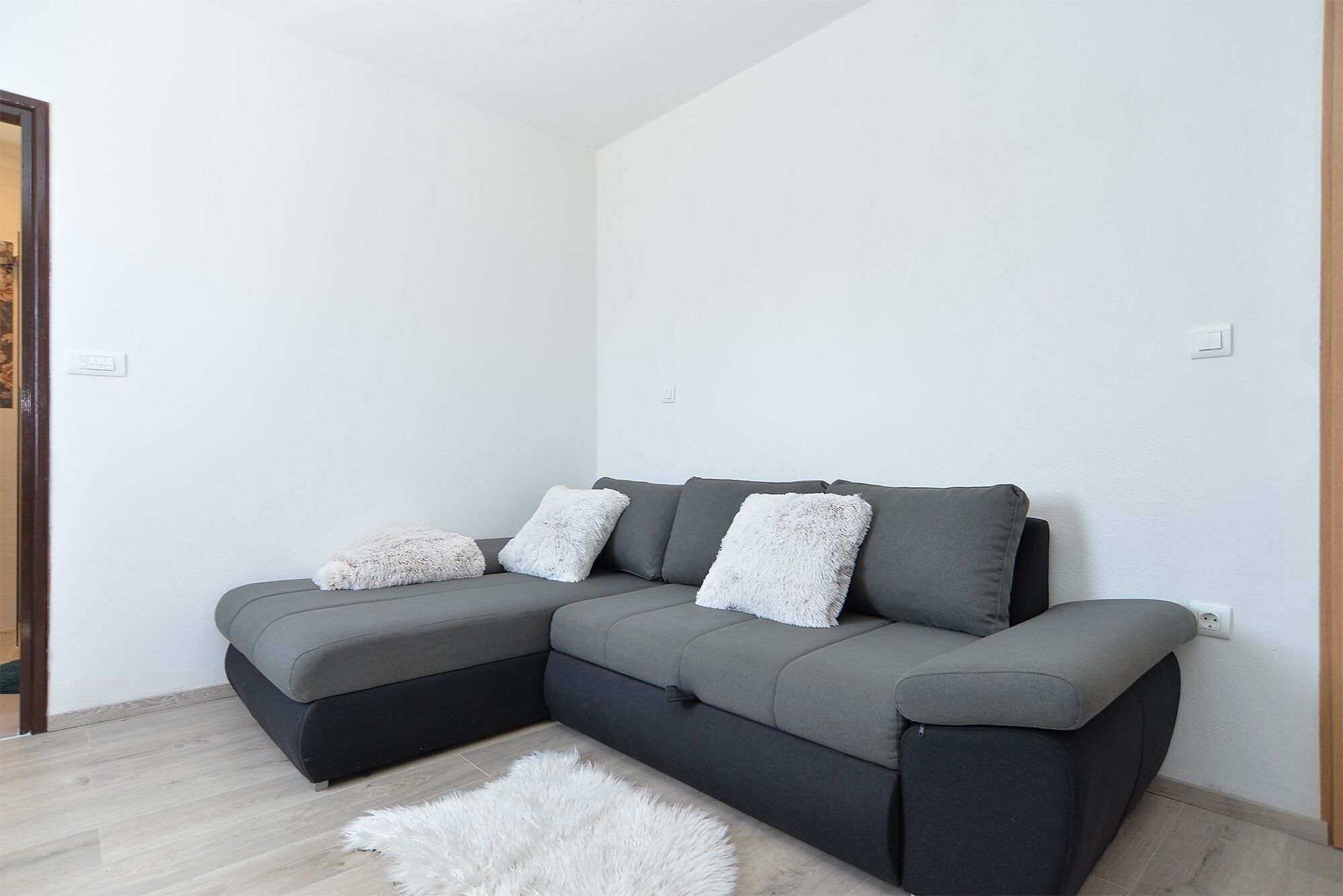 A grey sectional sofa with three white fluffy cushions against a white wall, a wooden floor, and a white furry rug.