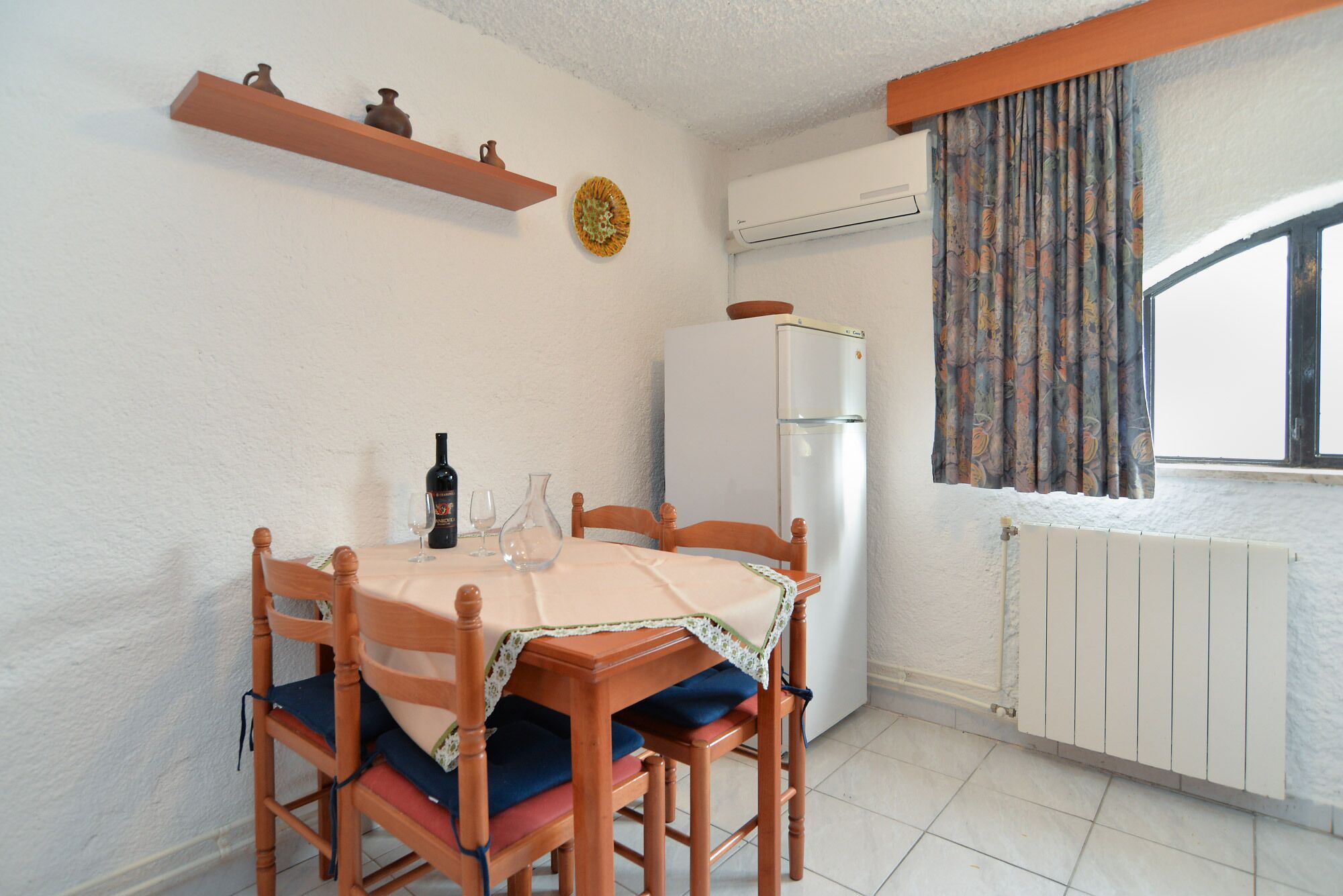 Un coin repas avec une table dressée pour deux personnes, un réfrigérateur, une fenêtre avec des rideaux et un radiateur.