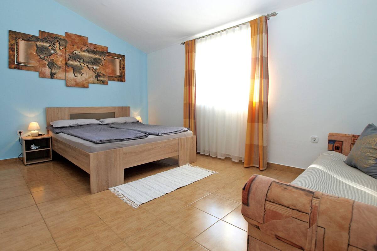 Une chambre à coucher avec un lit en bois, une table de chevet, une lampe fixée au mur et une grande fenêtre avec des rideaux.