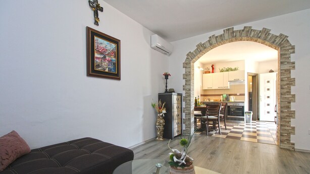 Un salón con sofá, una mesa de centro de vidrio y un jarrón decorativo. Se ve una cocina a través de un arco.