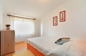 Ein Schlafzimmer mit einem Bett, einer Holzkommode, einem Fenster mit transparenten Vorhängen und zwei gerahmten Bildern an der Wand.