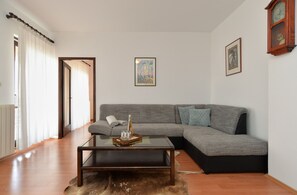 A living room with a grey sectional sofa, a glass coffee table, and a fur rug. There are two framed pictures on the wall, a wooden clock, and a door with curtains. - Apartment 1803 (Vodnjan)