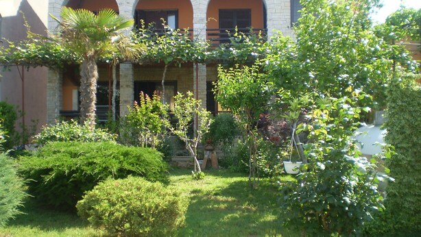 A courtyard with a brick building featuring arched windows and a well-maintained garden with various plants and a palm tree. - Apartment 1803 (Vodnjan)