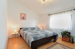 A bedroom with a bed, bedside lamps, a framed picture, a window with curtains, and a potted plant. - Apartment 1803 (Vodnjan)