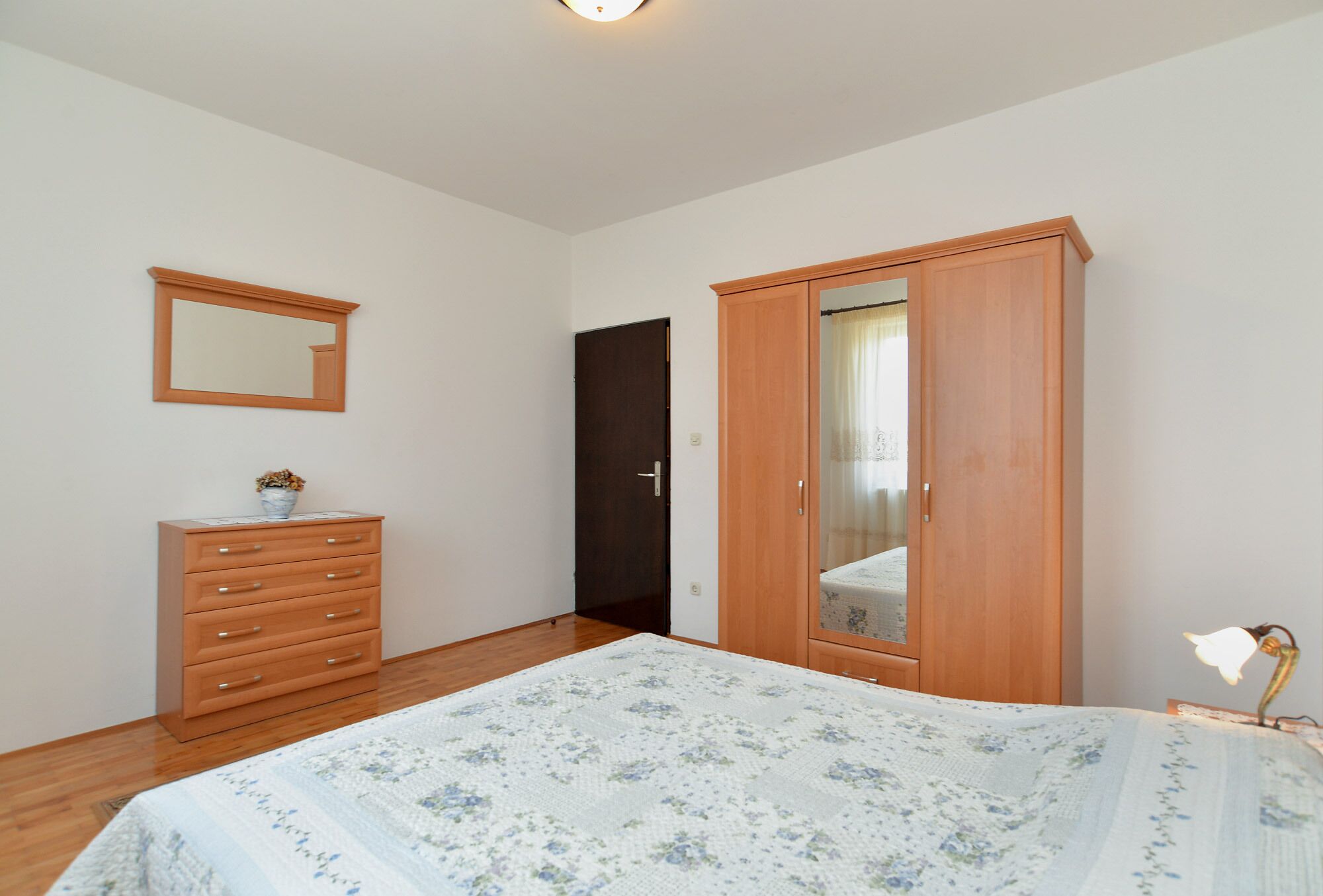 A bedroom with a wooden dresser, a large wardrobe, a bed with a floral bedspread, and a mirror on the wall.