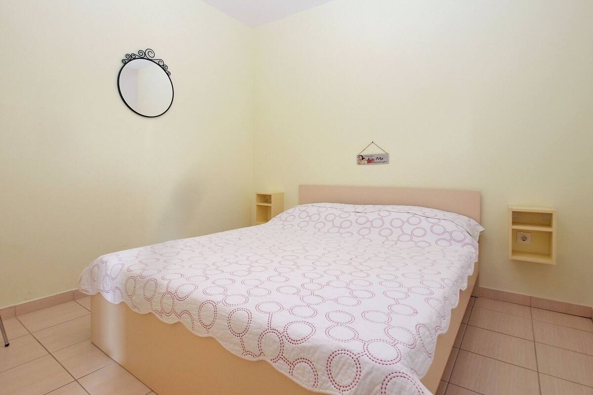 A bedroom with a single bed, a bedside table, a nightstand, a wall-mounted mirror, and a small shelf with a sign.