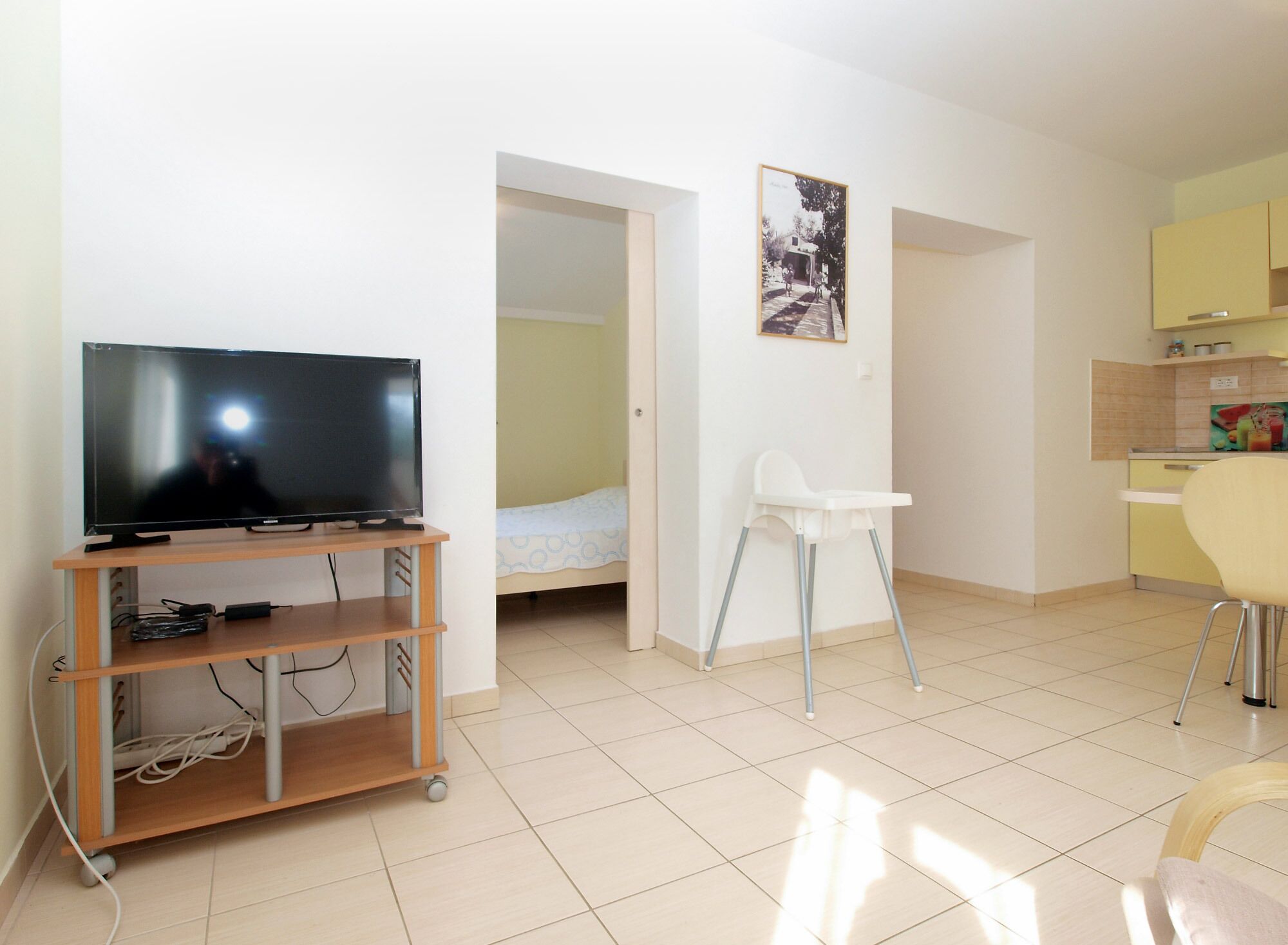Een knus woongedeelte met een televisie op een houten standaard, een witte stoel, een eethoek met een geel kastje, en een bed in de aangrenzende kamer.