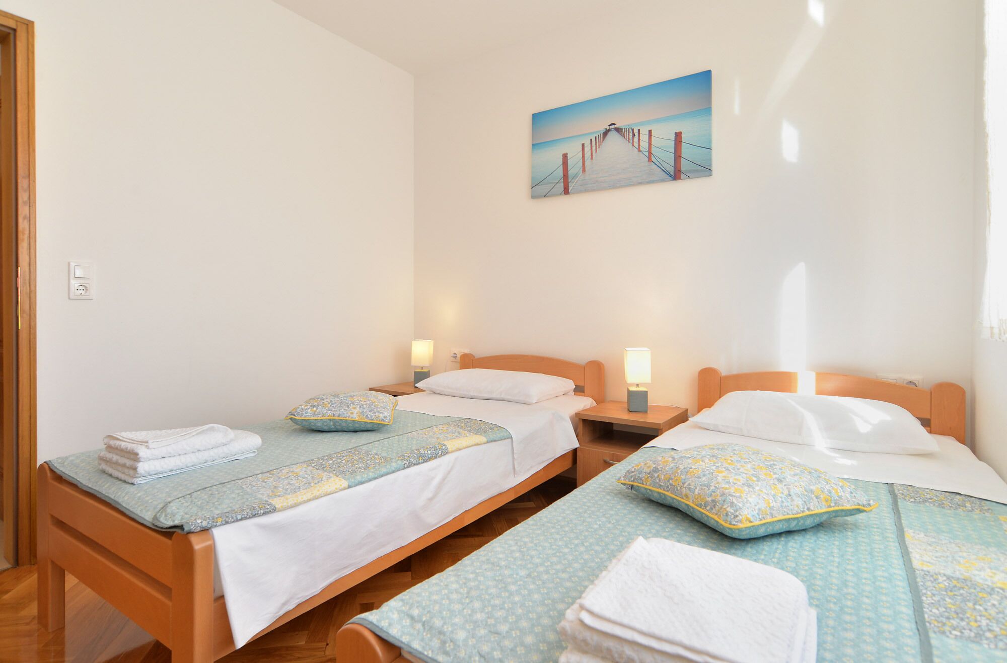 Een hotelkamer met twee eenpersoonsbedden, elk met een kussen en een bedlampje. Daarboven hangt een schilderij van een pier aan de muur.
