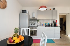 Une cuisine moderne avec des armoires blanches, une cuisinière en acier inoxydable et un four à micro-ondes. Un panier de fruits est posé sur la table à manger, et un luminaire suspendu se trouve au-dessus du comptoir.