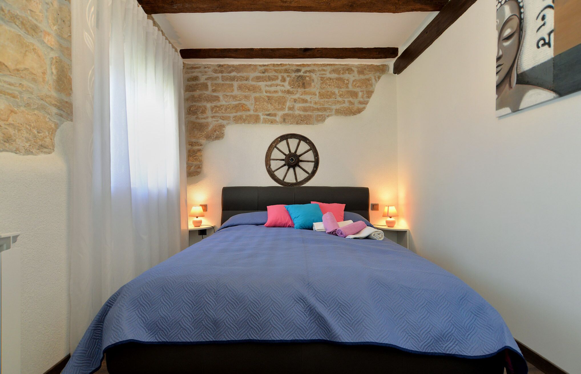 A bedroom with a stone wall, a wooden ceiling beam, a bed with a blue bedspread and colorful pillows, and two bedside lamps.