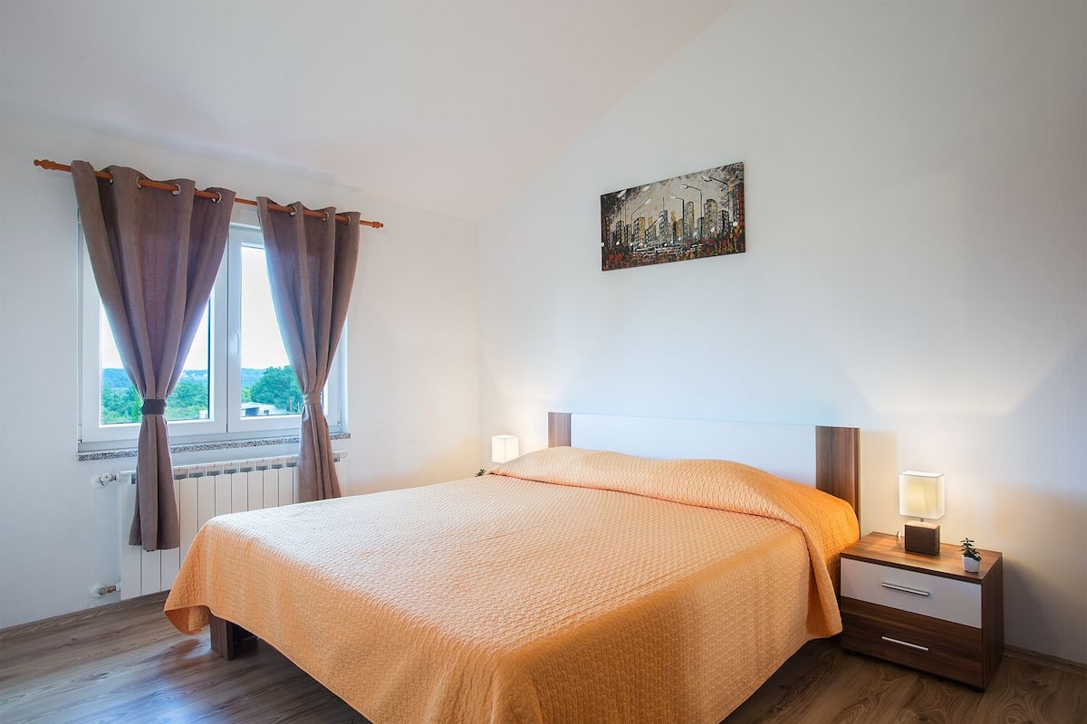A bedroom with a bed, a nightstand, a lamp, a window with curtains, and a painting on the wall.