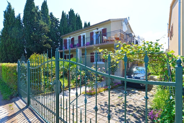Ein zweigeschossiges Haus mit Balkon, umgeben von einem Garten und einem Metallzaun.
