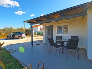 A covered outdoor patio with a table and chairs, a car parked outside, and a building with a window and door. - House 1818 (Pula)