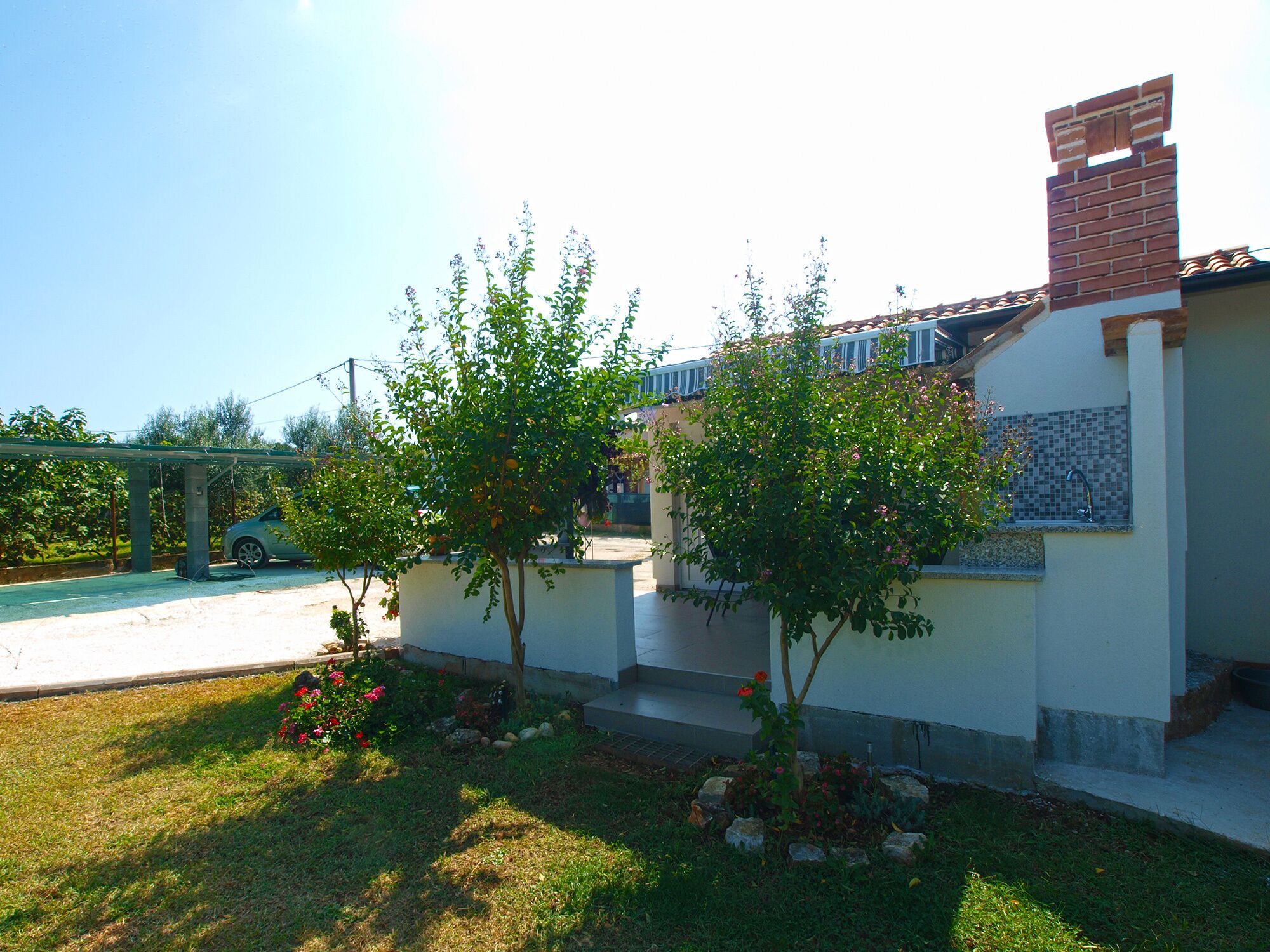 Un edificio residencial con fachada blanca, una chimenea de ladrillo y un pequeño jardín con plantas en flor y un árbol.