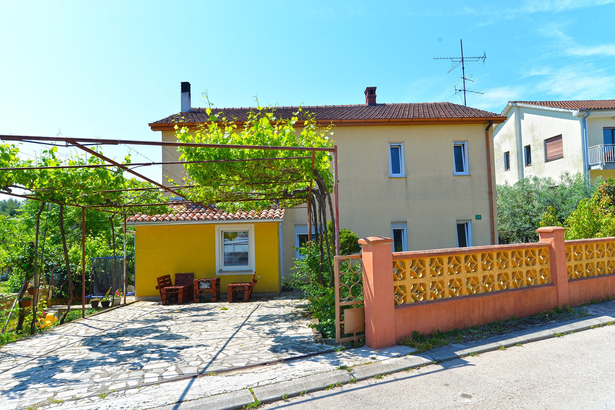 A yellow building with a tiled roof, a small porch with chairs, and a stone-paved entrance.