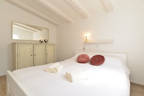 A neatly made bed with rolled towels and two pillows, a white dresser with a mirror, and a wall-mounted lamp. - House 1833 (Rovinj)