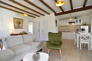 A living room with a sofa, a green chair, and a small table with a vase. There is a kitchen in the background with white cabinets and a dining area. - House 1833 (Rovinj)