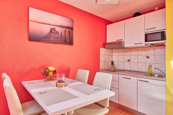 A compact kitchen with white cabinets, a microwave, and a sink. There is a dining table with chairs and a fruit bowl. A painting of a pier is on the wall.
