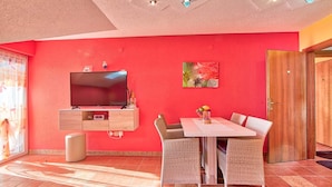 Une pièce avec un mur rouge, un téléviseur à écran plat fixé au-dessus d’un meuble en bois, des chaises en osier disposées autour d’une table blanche et une porte en bois sur la droite.
