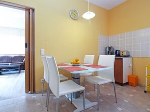 Comedor con mesa de vidrio, sillas blancas y frutero. Zona de cocina con cafetera y tostadora.