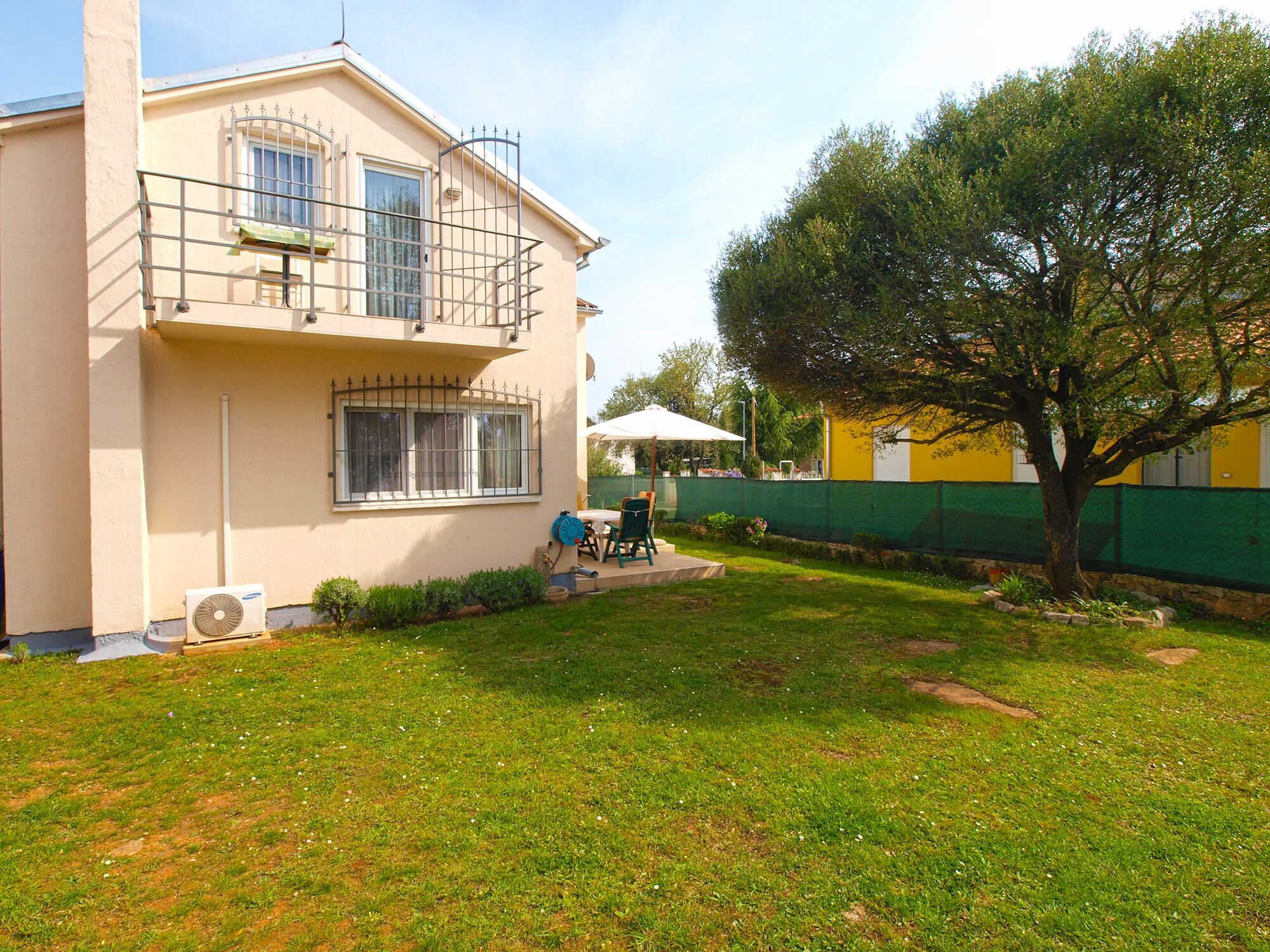 A two-story house with a balcony, a small patio with a table and chairs, and a tree in the yard.