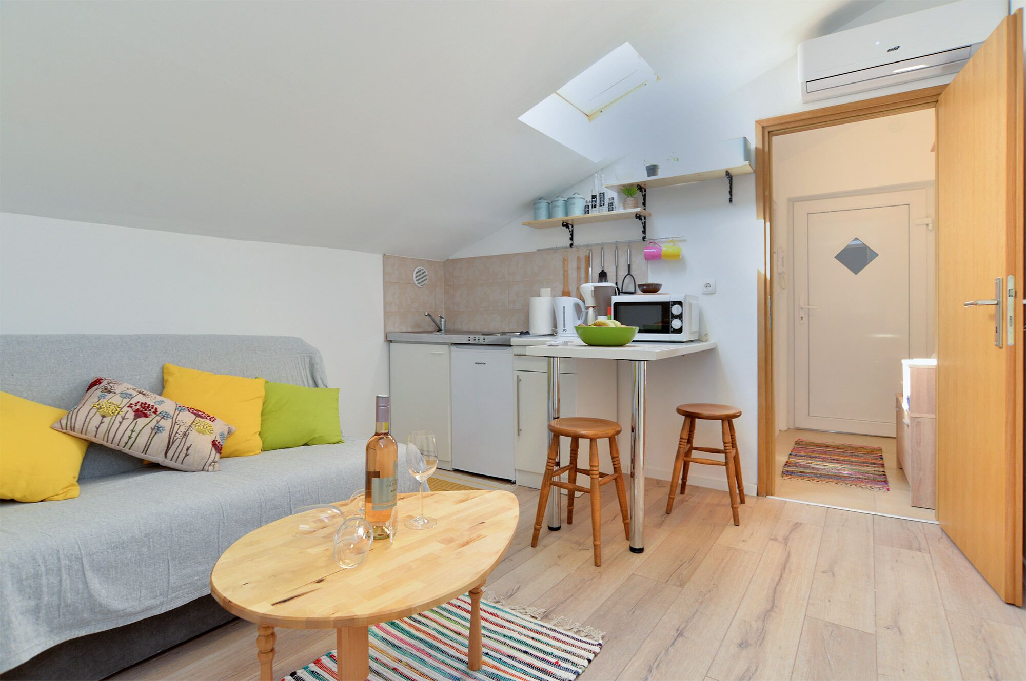 A compact living space with a kitchenette, a sofa with colorful cushions, a dining table, and a small stool.