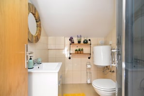 Une salle de bain avec un lavabo blanc, des toilettes et un espace douche. On y trouve des étagères avec des articles de toilette et un miroir au-dessus du lavabo.