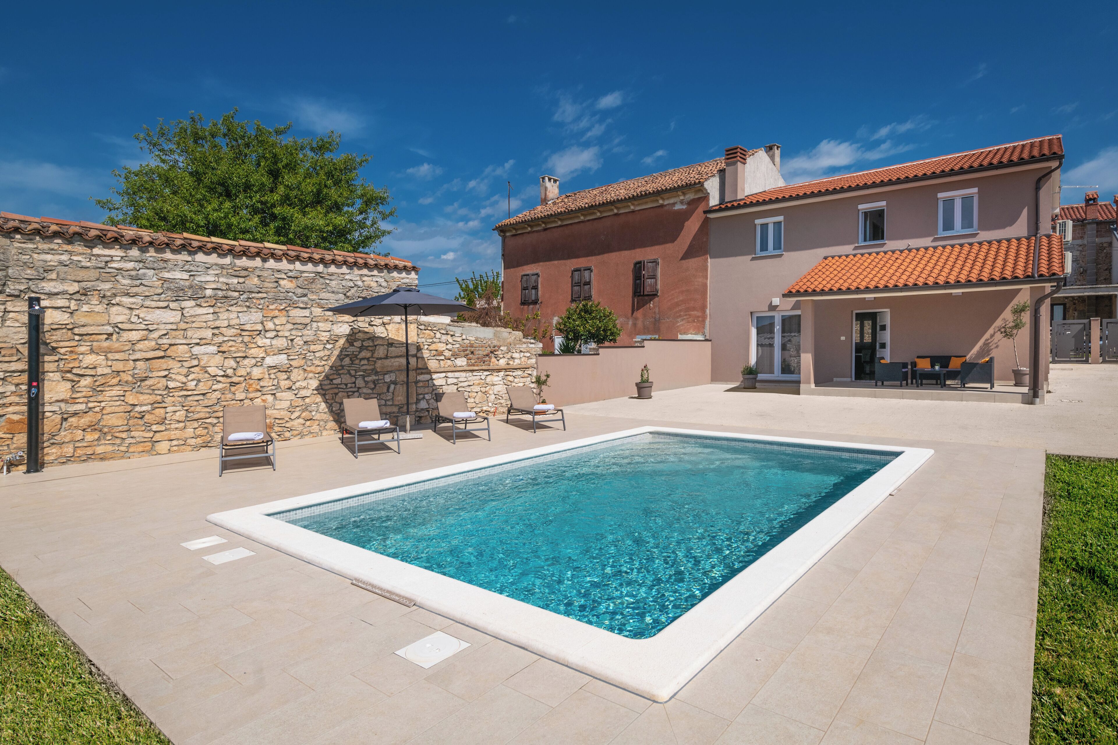 Una piscina con muro de piedra y sillones de descanso, rodeada de césped cuidado y un edificio con tejado de baldosas.