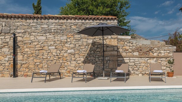 A stone wall with a tiled roof, a pool with clear blue water, and three lounge chairs with a black umbrella. - House 1855 (Liznjan)