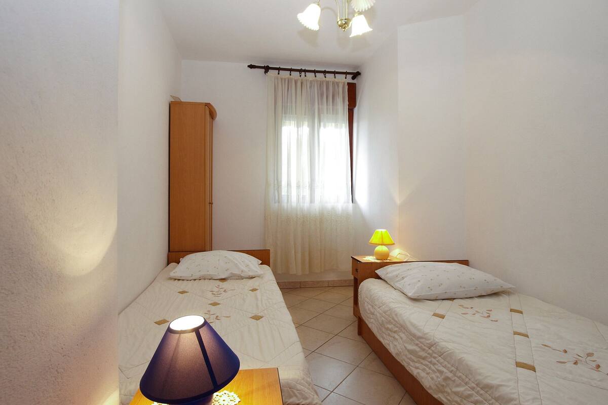 A small, well-lit bedroom with two single beds, a wooden wardrobe, a bedside table with a lamp, and a window with sheer curtains.