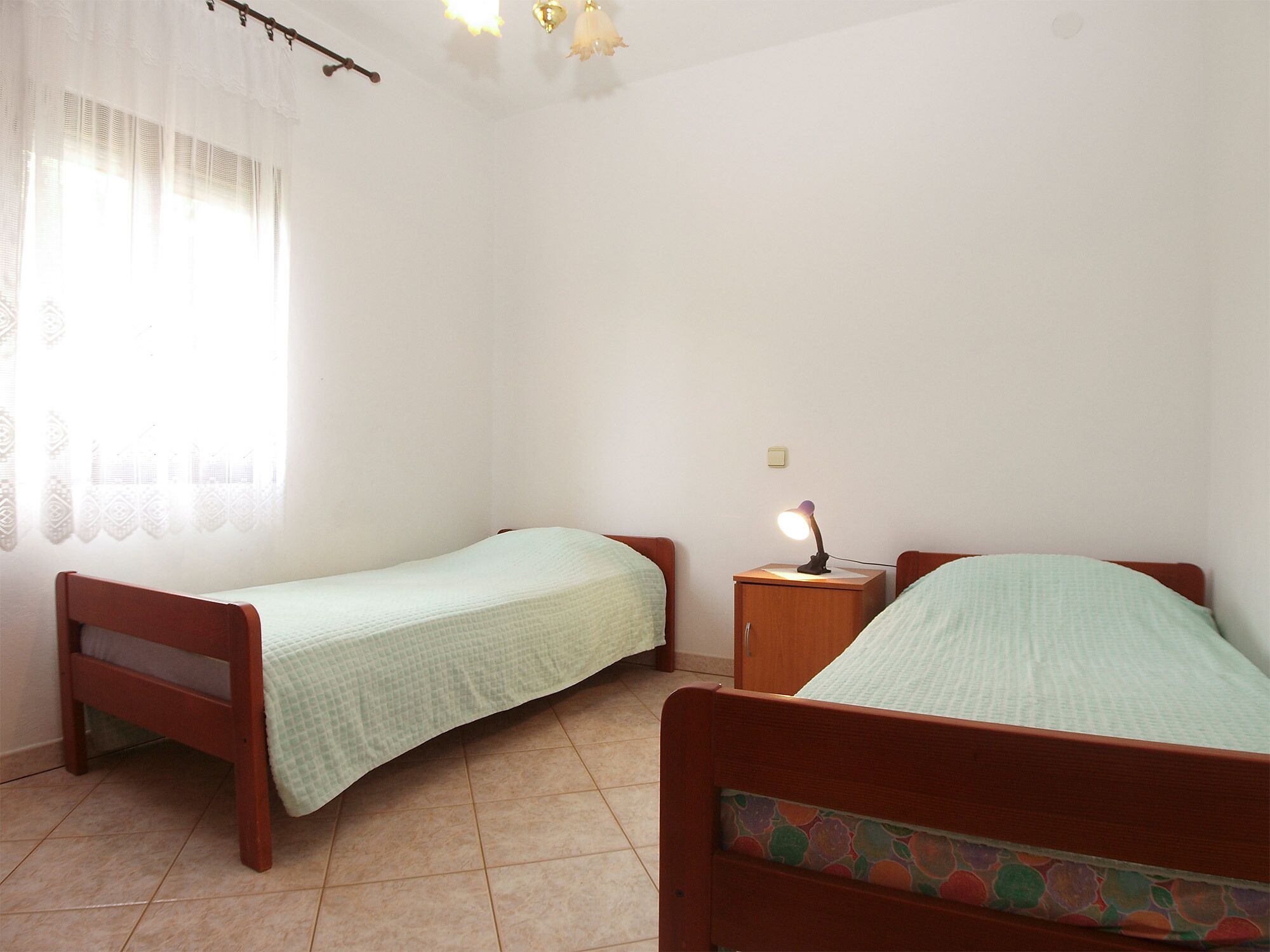 Une petite chambre propre, avec deux lits simples, une table de chevet avec une lampe et une fenêtre ornée de rideaux légers.