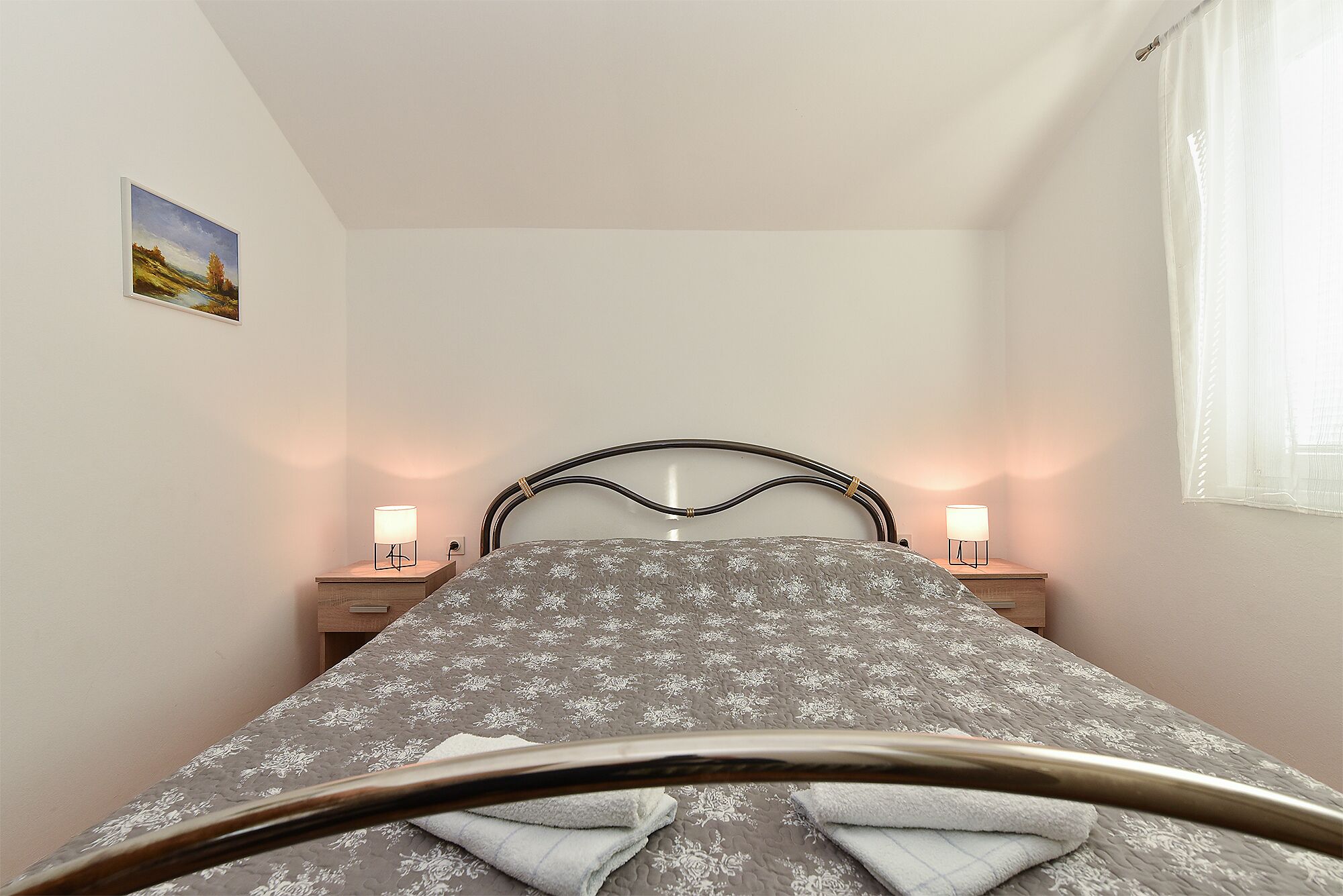 Un dormitorio con una cama, dos mesitas de noche con lámparas, un cuadro en la pared y un ventanal con cortinas.