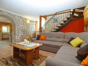 A living room with a stone wall, a wooden coffee table, a grey sofa with orange and yellow cushions, and a wooden staircase. - Apartment 1862 (Zminj)