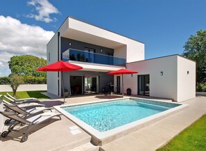 Une maison moderne à deux étages dotée d’une piscine, d’un espace salon extérieur et de parasols rouges.