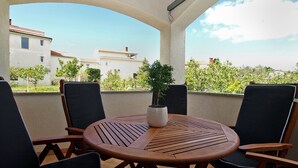 A covered outdoor seating area with a round wooden table and four chairs, a potted plant, and a view of buildings and trees. - Apartment 1868 (Pula)