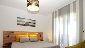 A bedroom with a wooden headboard, a bed with striped bedding, two bedside lamps, a floor lamp, a green armchair, a large window with curtains, and a painting on the wall. - Apartment 1868 (Pula)